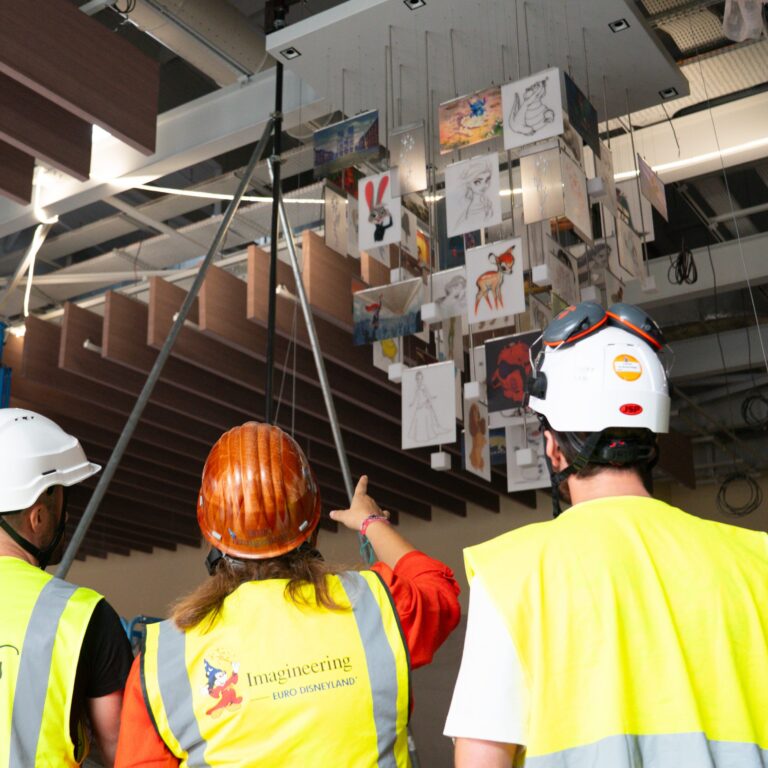 Three members of the technical team, wearing hard hats and safety vests, observe the installation of a chandelier featuring suspended Disney character sketches and illustrations.