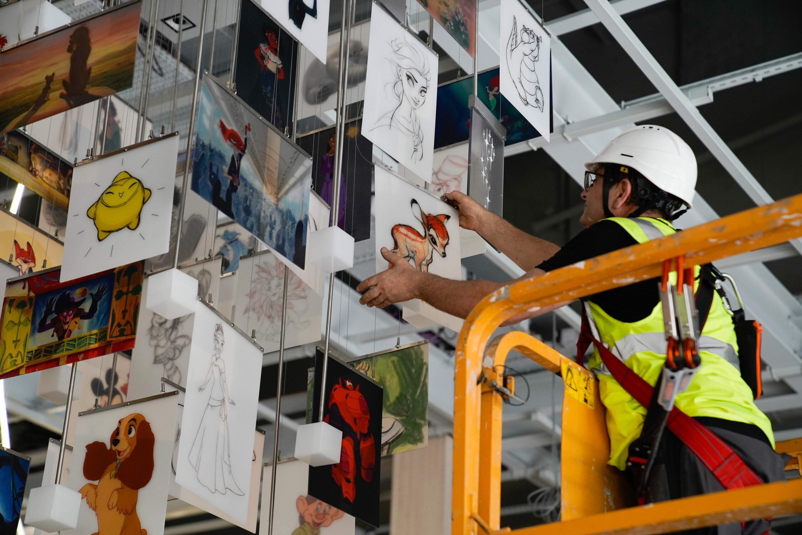 A technician wearing a hard hat and safety harness adjusts suspended panels featuring Disney characters on a chandelier from an aerial work platform.